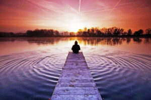 A person sitting on a dock, surroundedb7 calm water and a beautiful sunset.